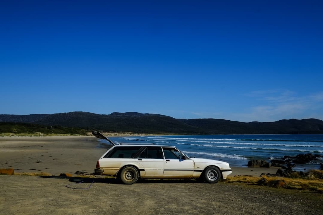 White rental car parked on a scenic coastal road in Australia overlooking a turquoise ocean and sandy beach, representing che