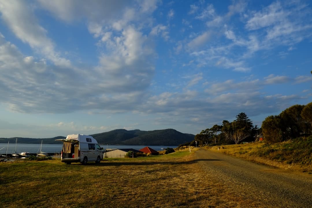 A modern white SUV drives along a scenic coastal road in Tasmania, with ocean views and green hills under a partly cloudy sky